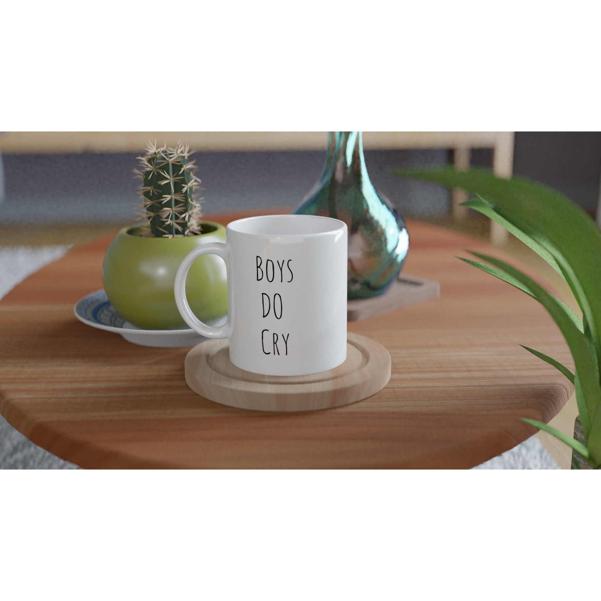 Boys Do Cry White 11oz Ceramic Mug on a wooden table, surrounded by plants.